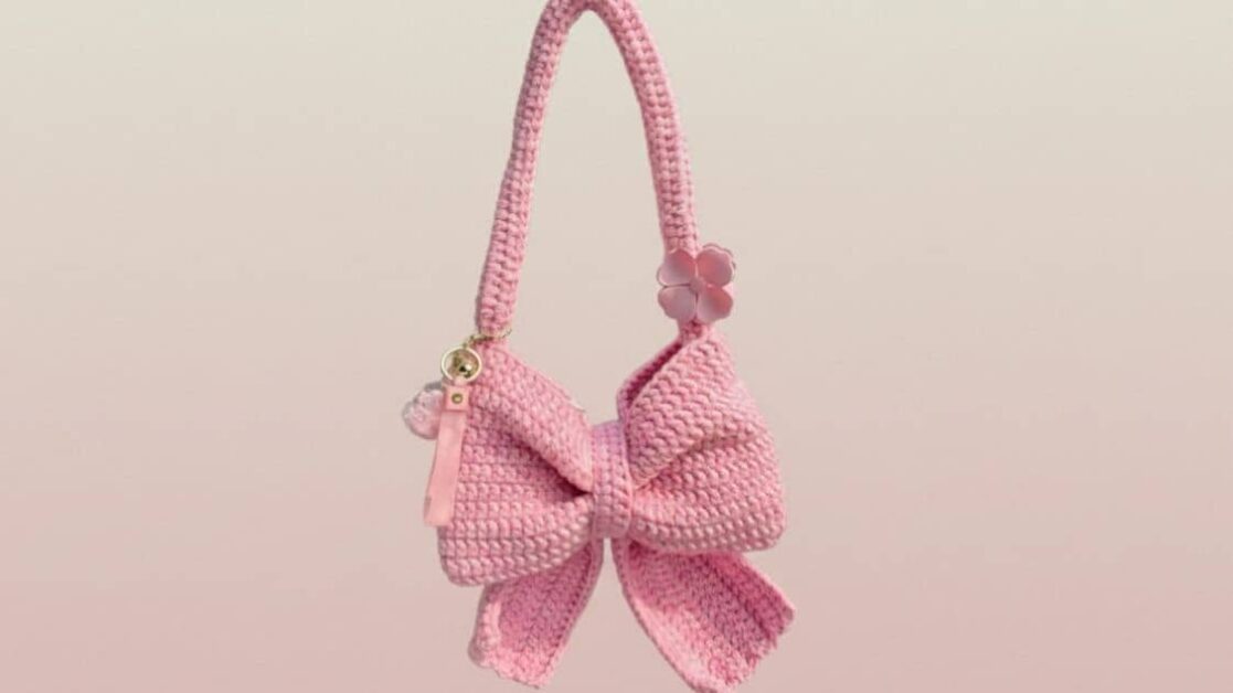 Bolsa de crochê rosa em formato de laço pendurada, com detalhes delicados como uma flor e um chaveiro.