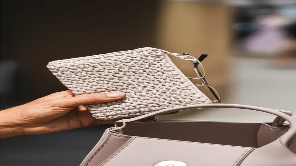 Mão segurando uma capa de óculos em crochê bege perto de uma bolsa aberta.