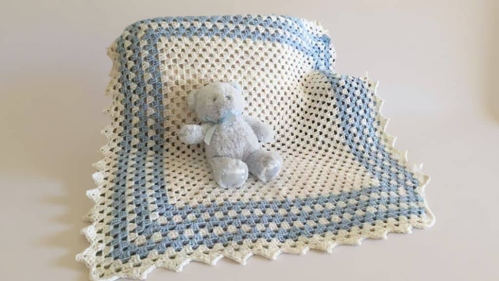 Manta de crochê para bebê em tons de azul e branco com um ursinho de pelúcia azul claro.