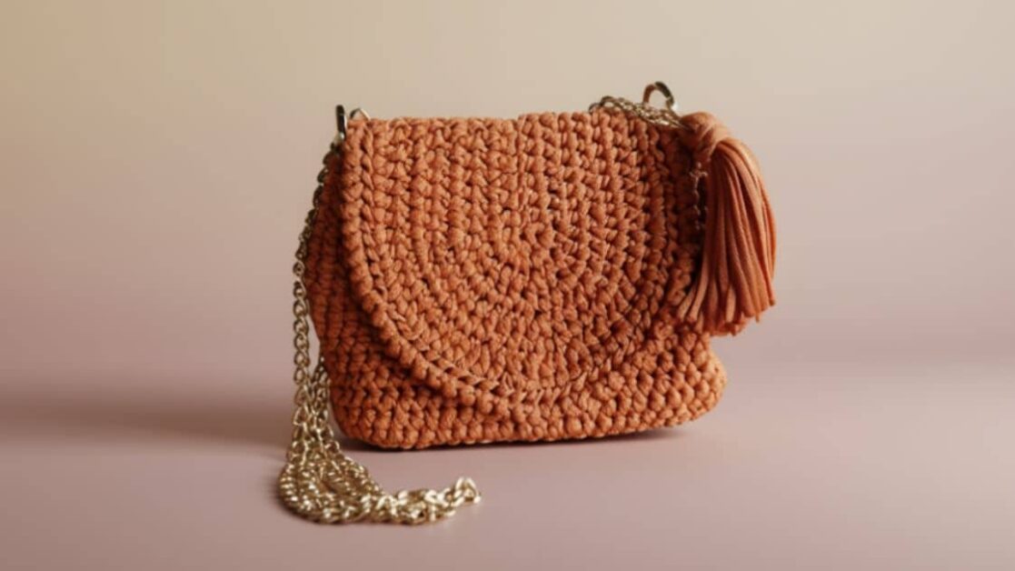 Bolsa de fio de malha cor de pêssego com alça de corrente dourada e detalhe de tassel.