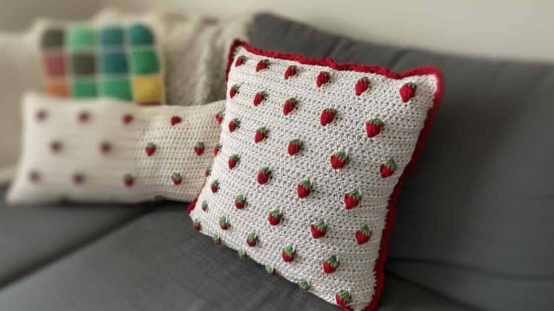 Almofada quadrada de crochê branca com morangos bordados e borda vermelha, em um sofá cinza.