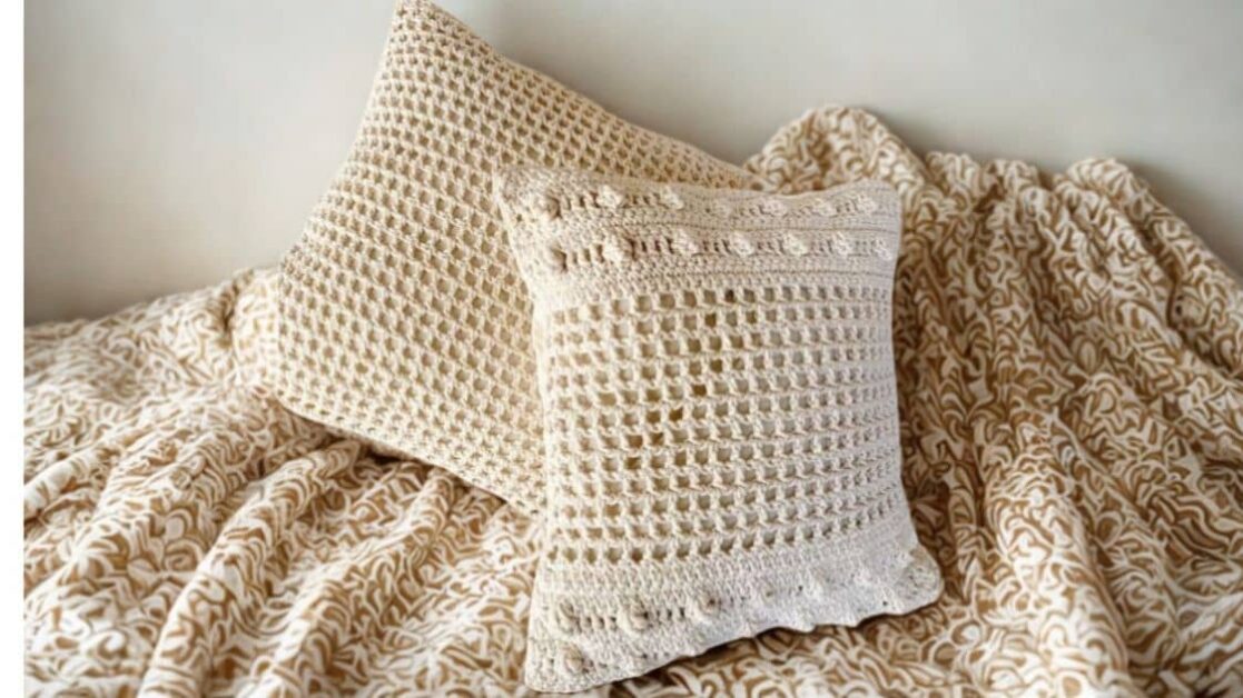 Duas almofadas de crochê bege em cima de uma manta com padrão abstrato bege e branco.