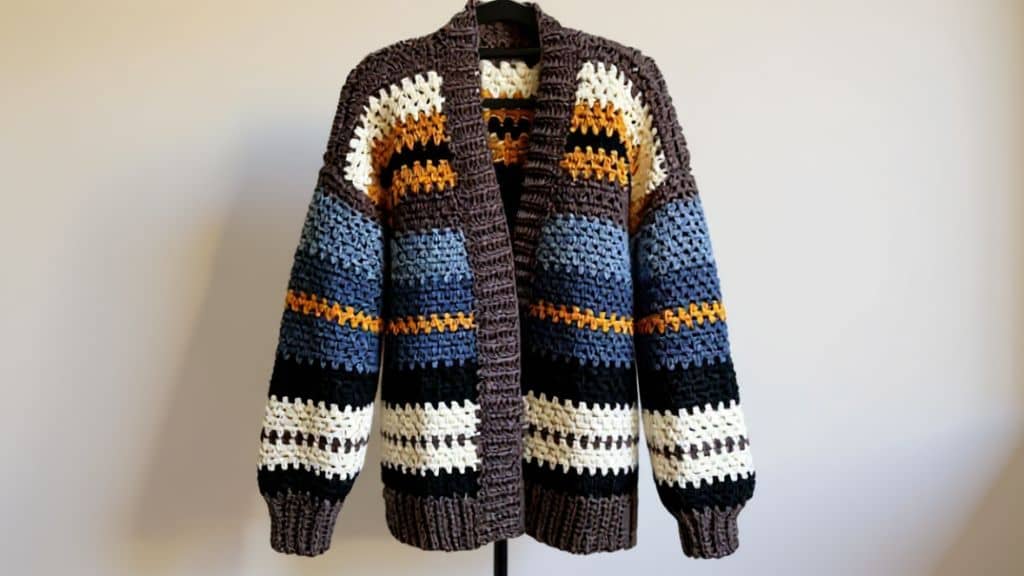 Casaco de crochê listrado em tons de marrom, azul, branco, preto e amarelo pendurado em um cabide.