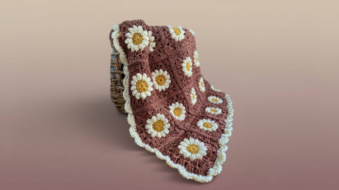 Manta de crochê granny square com motivo de margaridas, em tons de marrom, amarelo e branco, delicadamente disposta sobre um cesto de vime.