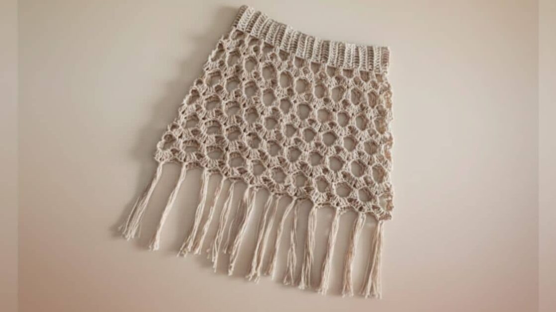 Saia de crochê bege com trama vazada e franjas longas na barra, ideal para o verão.