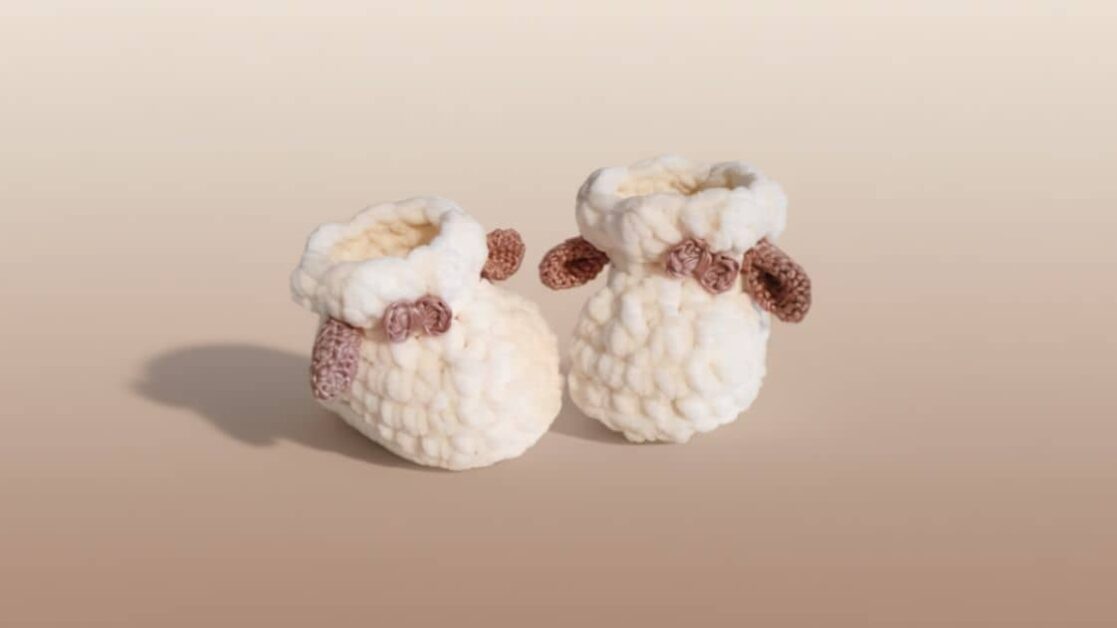 Dois sapatinhos de crochê brancos em formato de ovelha, com detalhes marrons nas orelhas e enfeites laterais.