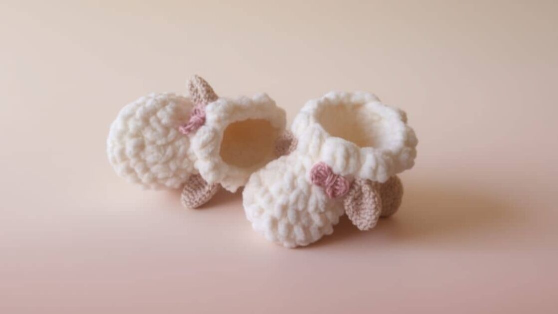 Par de sapatinhos de crochê em forma de ovelha, com detalhes em rosa e bege, sobre fundo rosa claro.