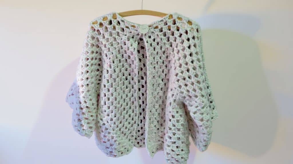 Cardigã de crochê branco pendurado em um cabide, com um único botão no topo.
