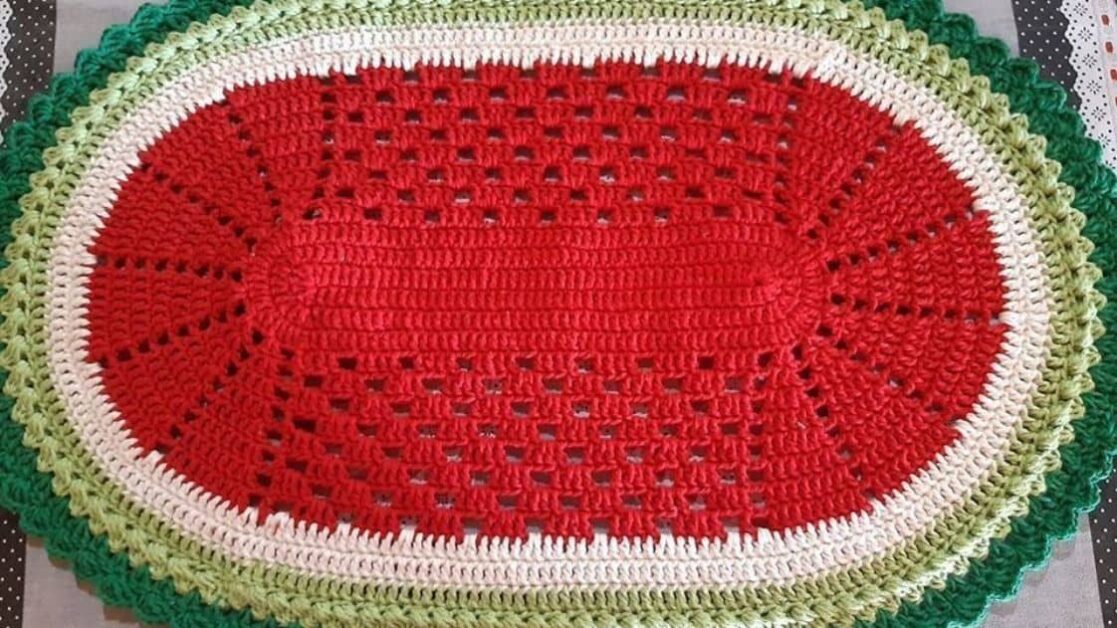 Tapete oval de crochê com design que remete a uma fatia de melancia, com bordas verdes e centro vermelho.