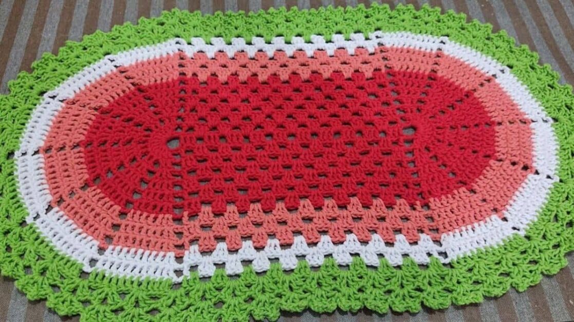 Tapete oval de crochê com design de melancia, mostrando as cores verde, branco, rosa e vermelho.