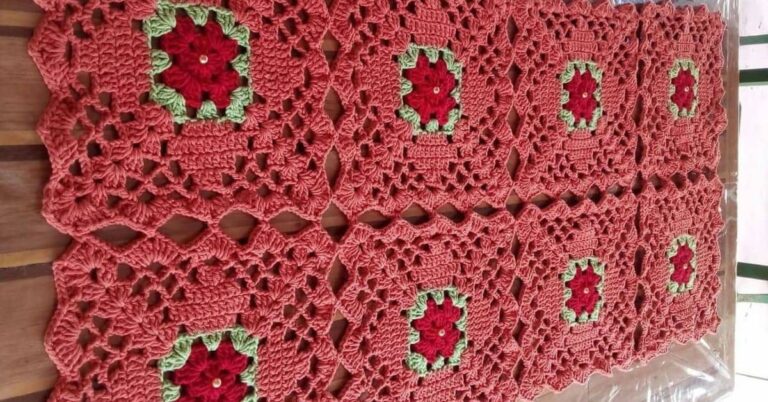 Trilho de mesa geométrico quadrado em crochê, com detalhes florais vermelhos e bordas verdes, sobre uma mesa de madeira.