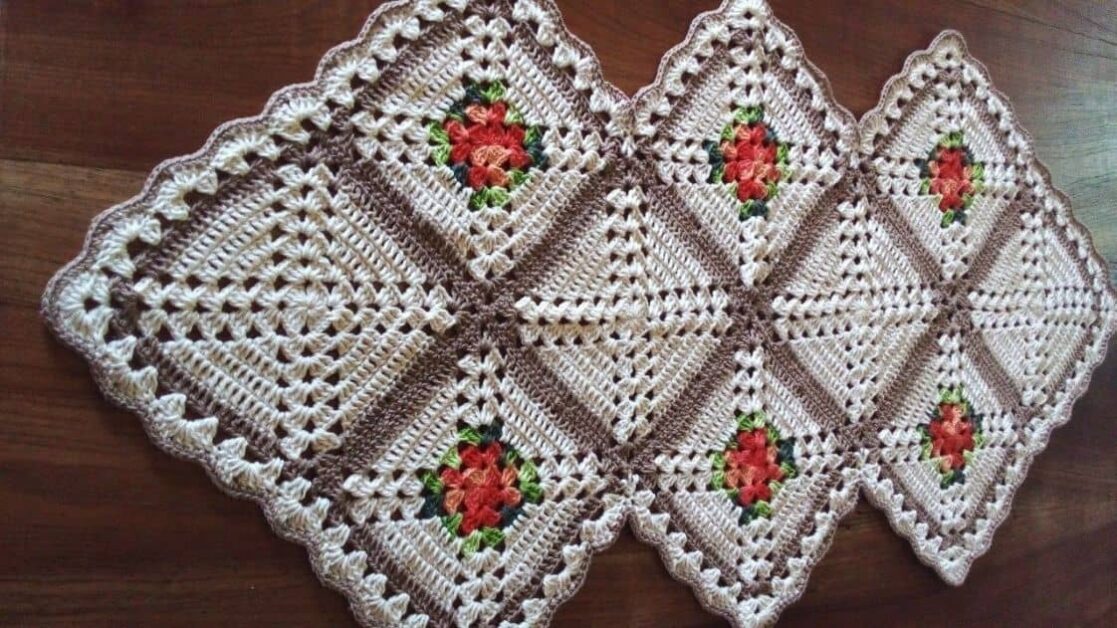 Trilho de mesa geométrico quadrado de crochê em tons de bege e marrom, com detalhes florais vermelhos e verdes.